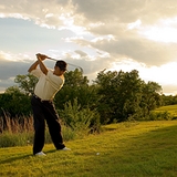 An outdoor sports photo of a golfer at the end of his swing. He is looking toward the setting sun and has scenic green grass, trees and satiny clouds around him.